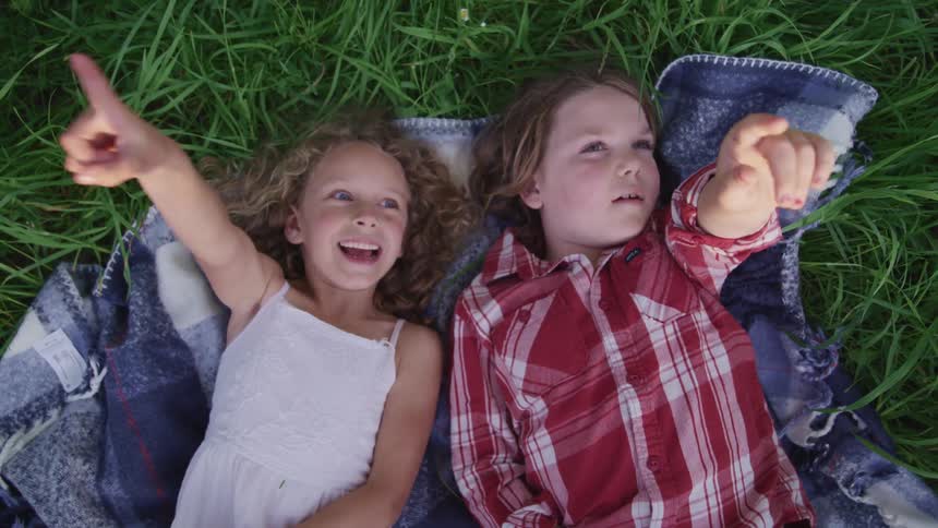 Two small children lying down on a blanket outside in the grass pointing upwards towards the sky