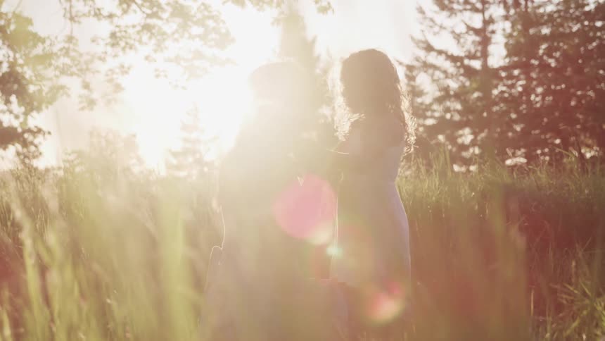 Mother and daughter hold hands share a kiss and laugh while standing in field with the sun shining