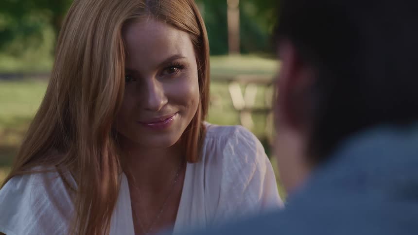 A young woman sitting at a table in a park looks up to smile at the man sitting across from her