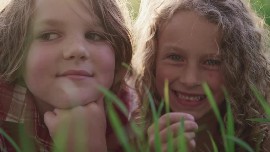 A young girl with long curly hair smiles as she poses lying down next to her brother behind tall grass on a lawn