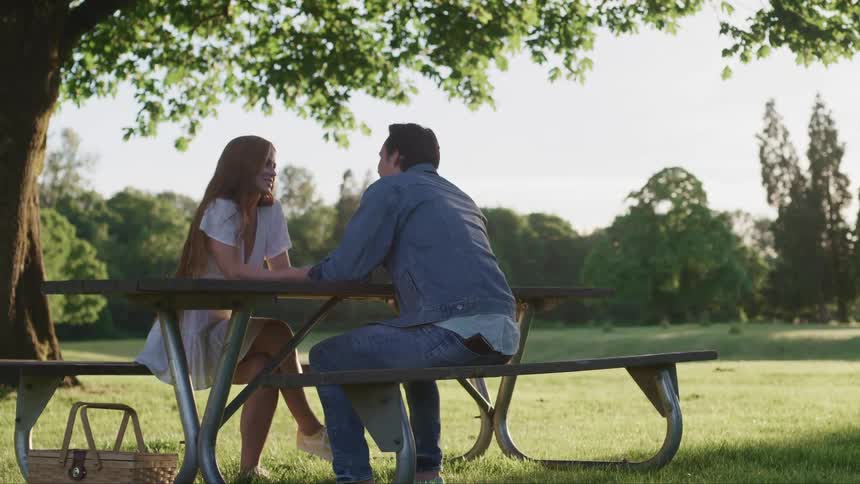 A man and woman sitting on a picnic table with trees the sky and clouds in the background