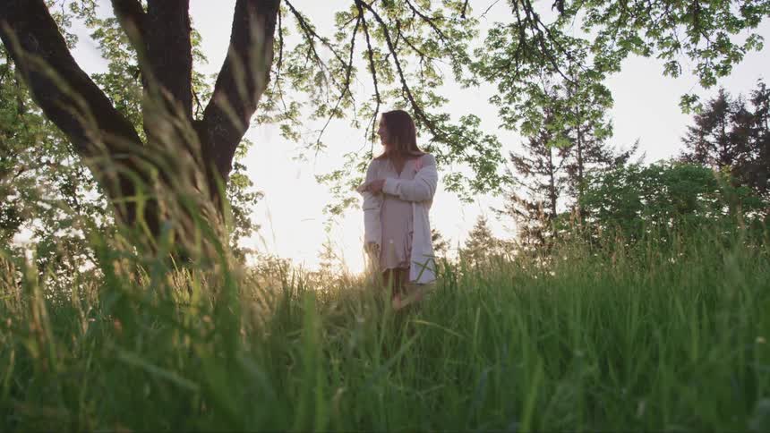 A woman wearing a white top stands under a tree on a field of long grass