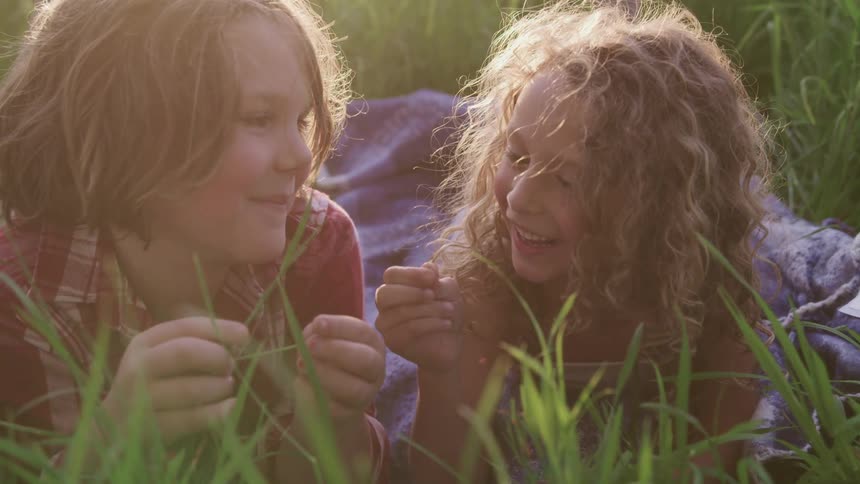 A girl and boy lie on a blanket on a field playing with the blades of grass