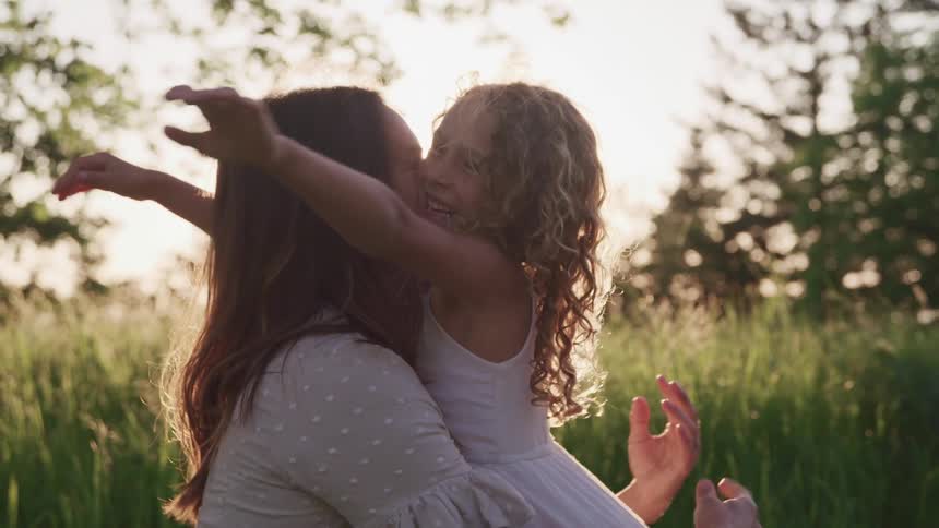 Mother kneeling down at level with her daughter at daytime gives her a hug and smiles outdoors in nature