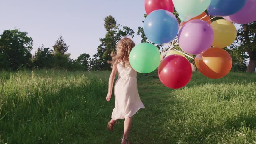 A little girl running on a grassy field with balloons with trees the sky and clouds in the background