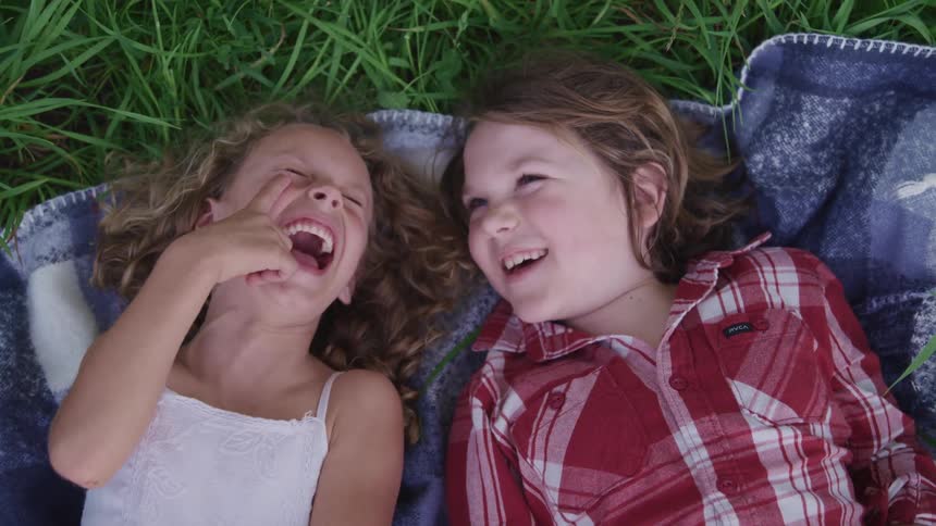 Two girls lie on backs on blanket on lawn smiling and laughing at each other