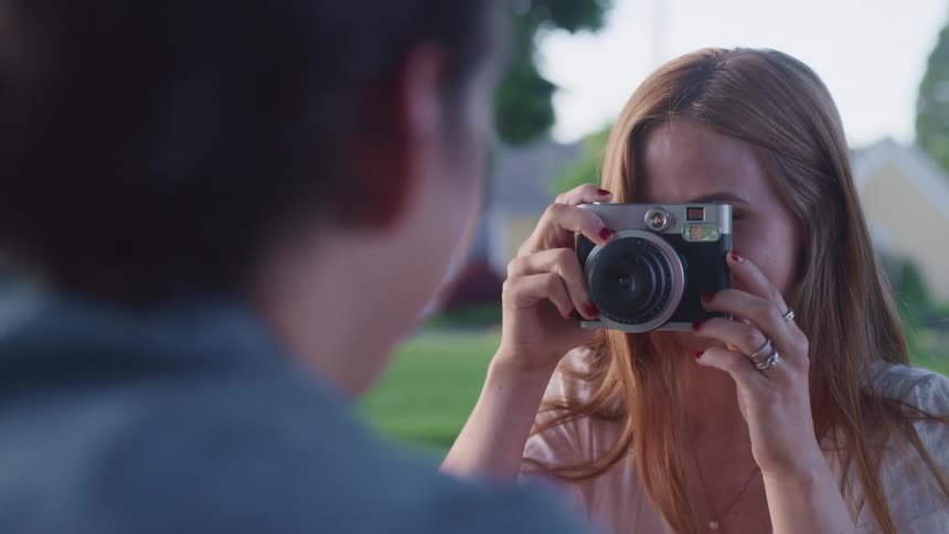 A woman takes a photo of a man with a camera and smiles outside sitting in a field of grass at daytime