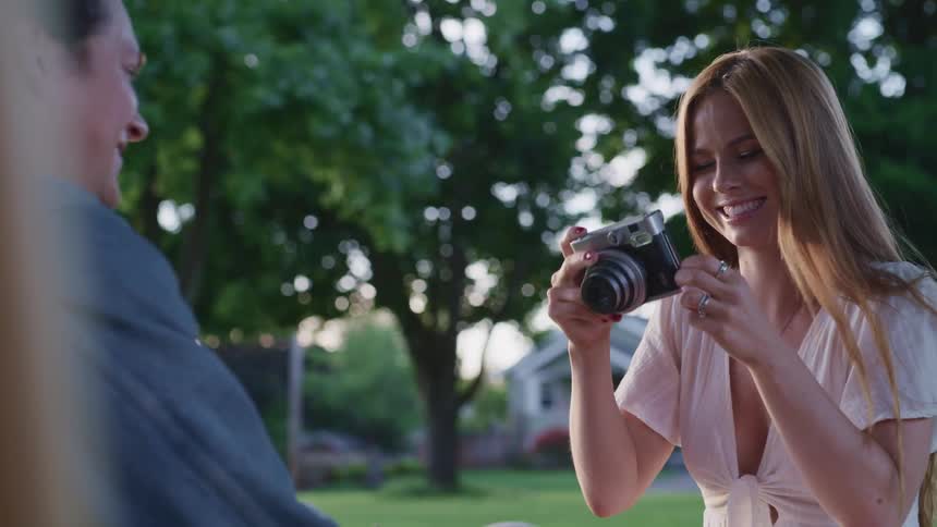 Young couple take photographs of each other with a vintage camera sitting over the grass in a park at evening