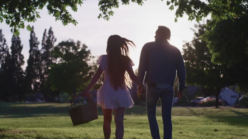 A woman carries a picnic basket as she walks through a park holding a mans hand