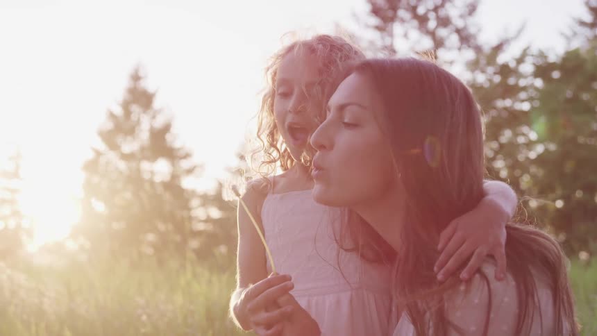 Mother and daughter blowing over a dandelion in a grass field at sunset