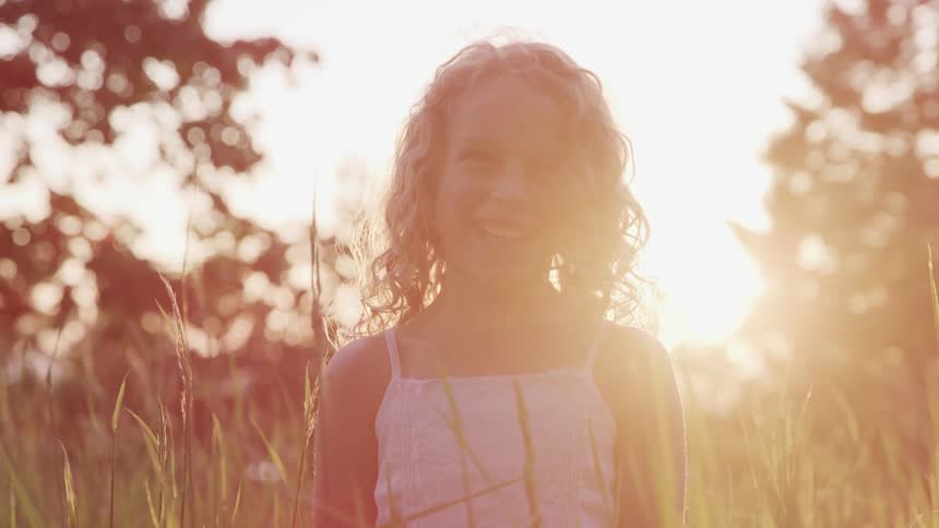 A little girl with curly hair standing in the middle of tall grass with trees and the sun behind her