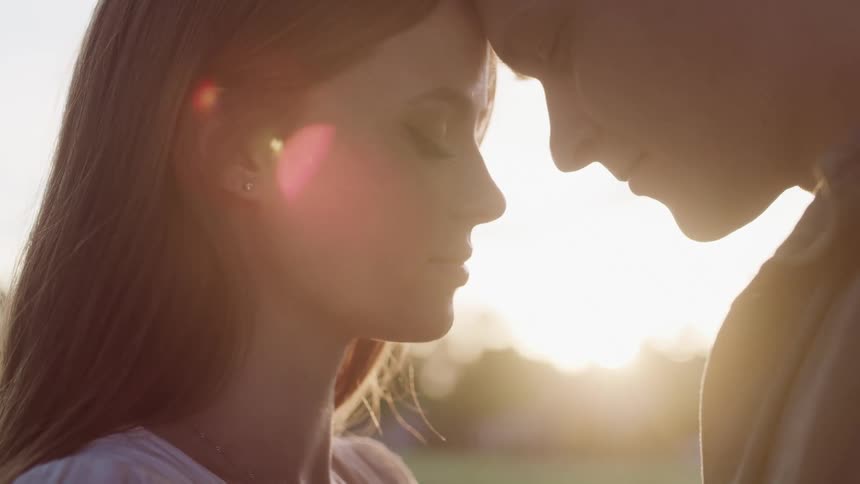 Man and a woman with their eyes closed and their heads pressed together while a flare of sun shines behind them