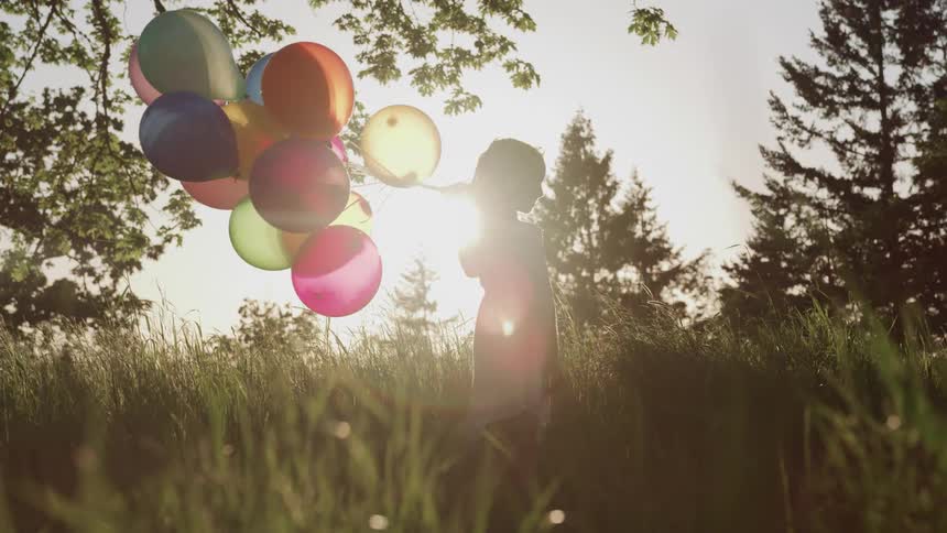 Young girl wearing a dress holding a bunch of colored balloons with the sun shining behind her and trees all around