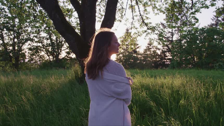 A woman standing in a field of grass in the evening slowly closes her eyes