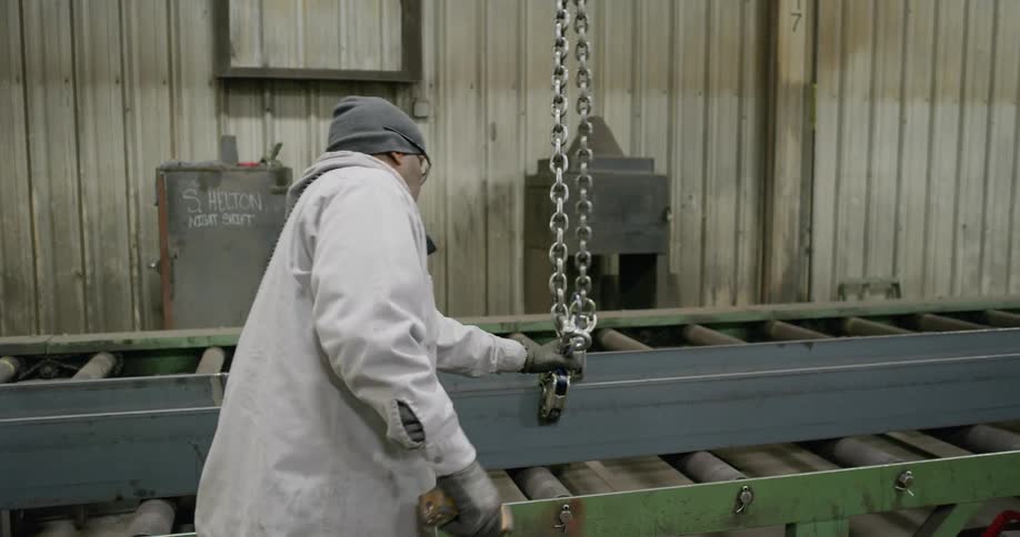 A worker in a factory moving a heavy chain along a long metal piece on a conveyor belt