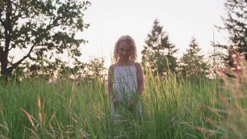You girl wearing white dress is standing in tall grass with trees in the distance