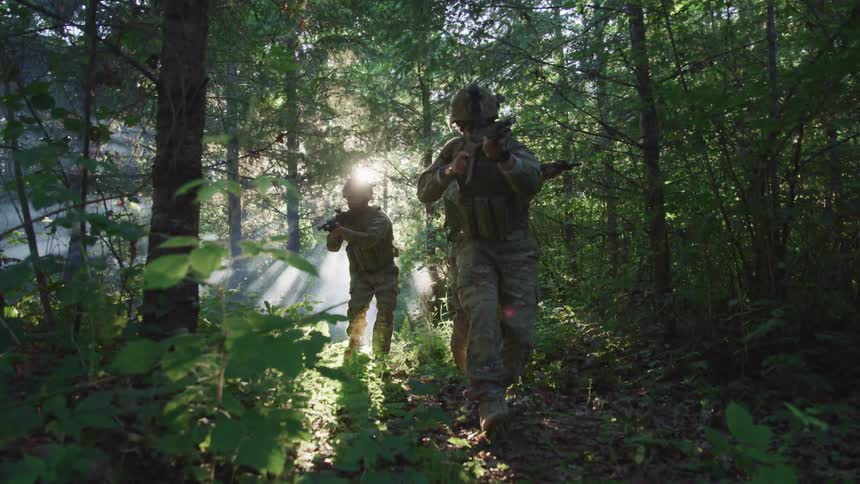 A small group of soldiers walk through a woodland with their firearms raised as sunlight shines through the foliage behind them
