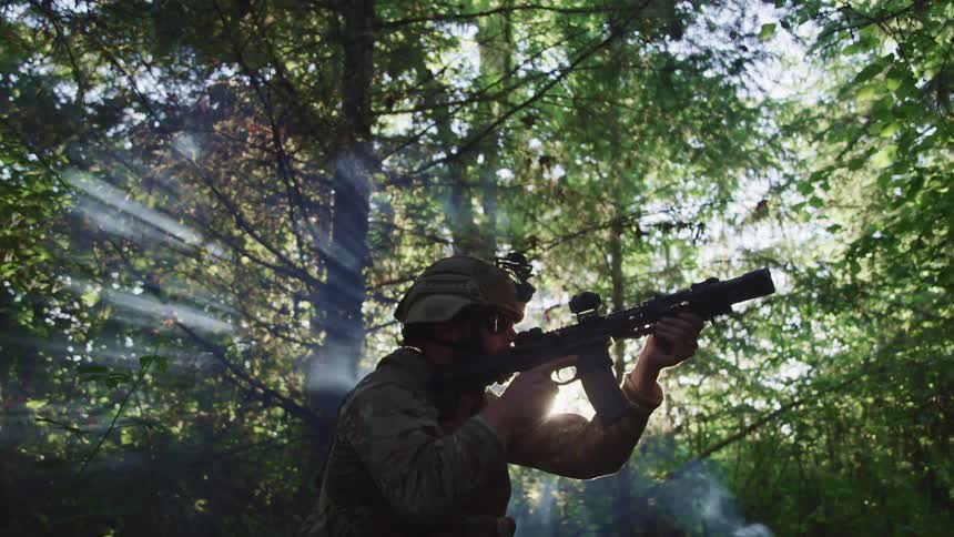 A soldier points his gun while standing in a forest during daylight hours