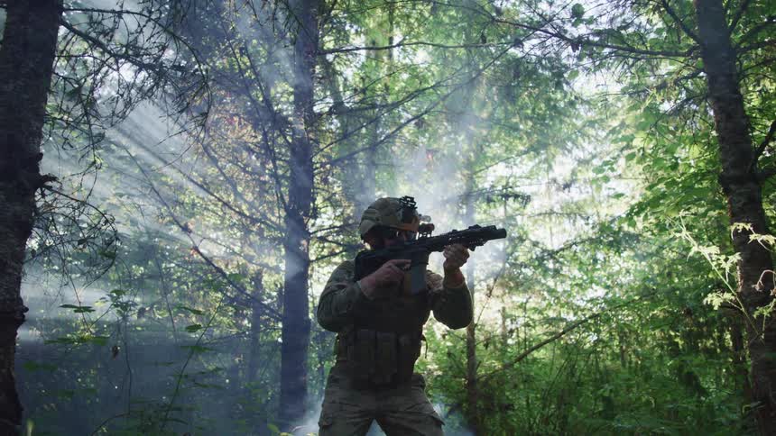 Solder in camouflage wearing a helmet is standing in the forest with gun ready and the sun is shining brightly from behind