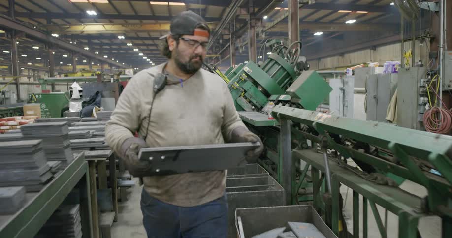 A worker moving a metal plate to a table with other metal plates in a factory with lots of different machinery