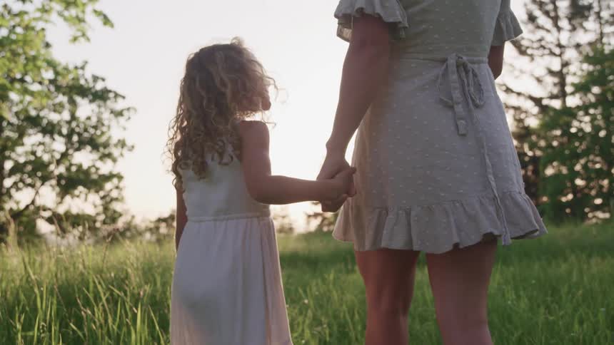 A girl turns to smile up at her mother as they walk hand in hand through a field during the day