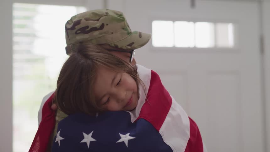Soldier hugs son wrapped in an american flag in a room at home