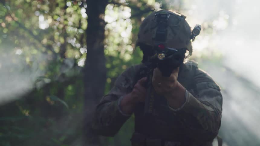 A soldier in camouflage stands and turns in a forest with his rifle drawn