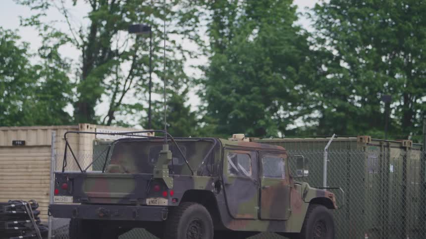 A camo painted jeep parked in front of a fence with trailers and shipping containers inside