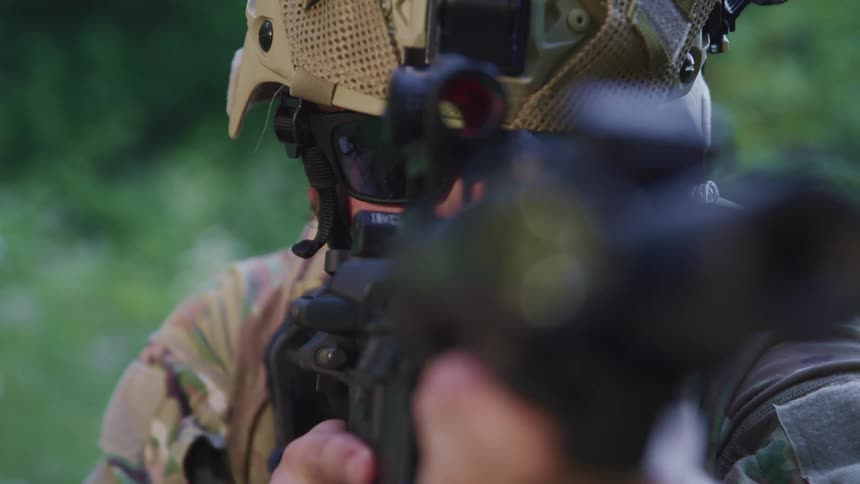 A man with a helmet and sunglasses pointing a rifle with trees and plants in the background