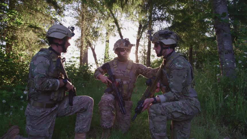 Three men in military gear holding guns down on one knee in a forest at daytime