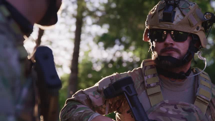 Two male soldiers with sunglasses weapons and helmets stand outdoors in greenery