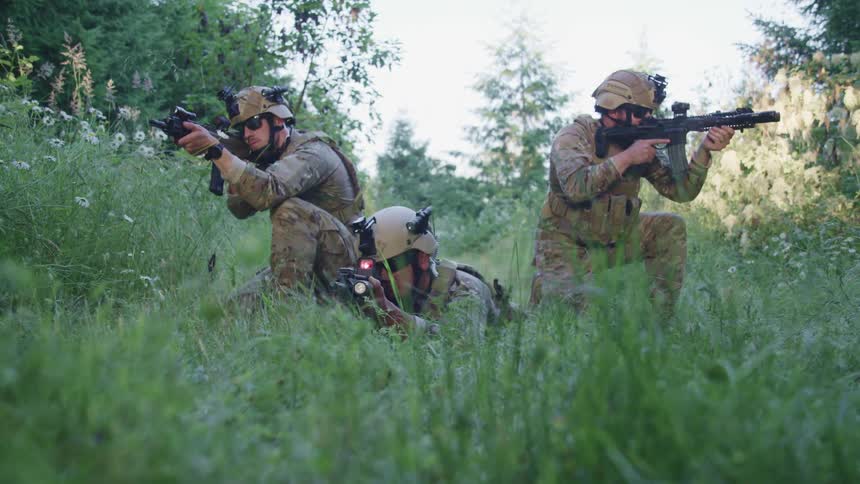 A trio of soldiers in combat uniform kneel with raised weapons in a field of tall grass during the day