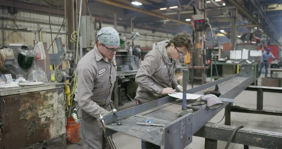 Men with goggles working in a factory as one uses a measuring tape