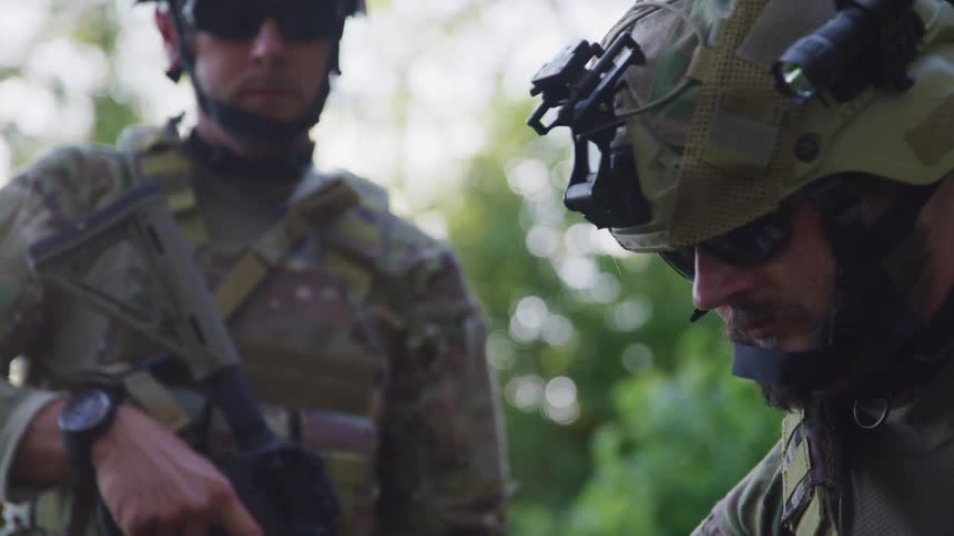 A soldier looking down next to another soldier standing up outside at daytime