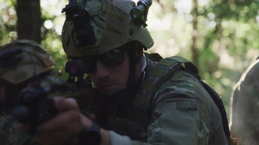 Soldier in a forest with other troops searches through the lens of a sniper rifle at daytime