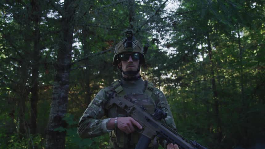 A soldier wearing combat gear holding a gun standing in the forest at evening