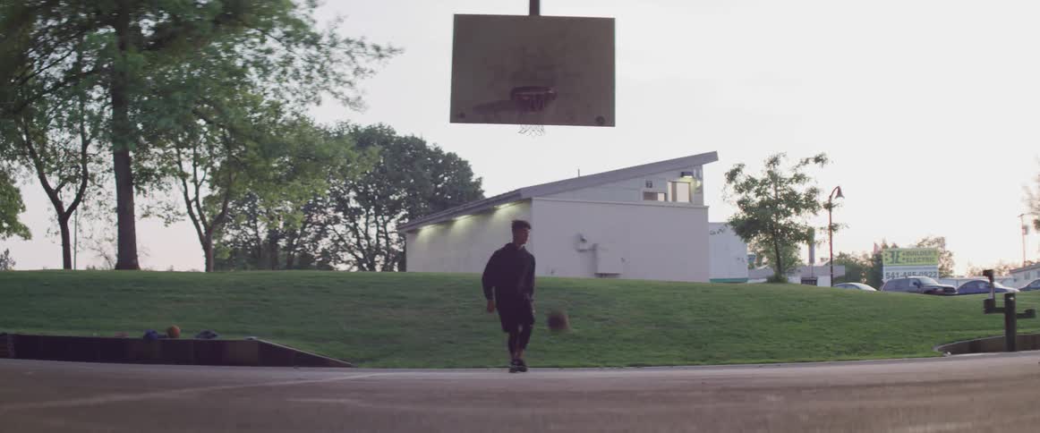 A young man playing basketball by himself in a park jumps and slams the basketball through the net