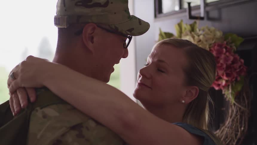 A woman hugs a soldier in the doorway and then they look at one another and smile