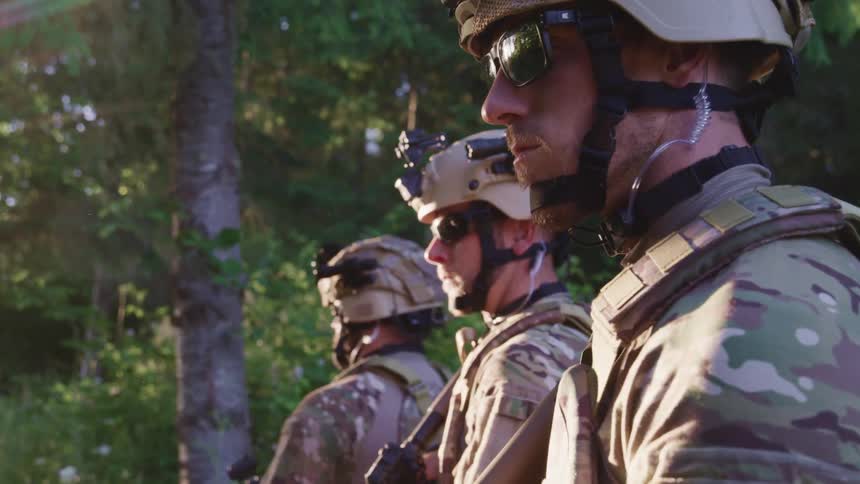 Three male soldiers stand outdoors near trees looking outwards during the day