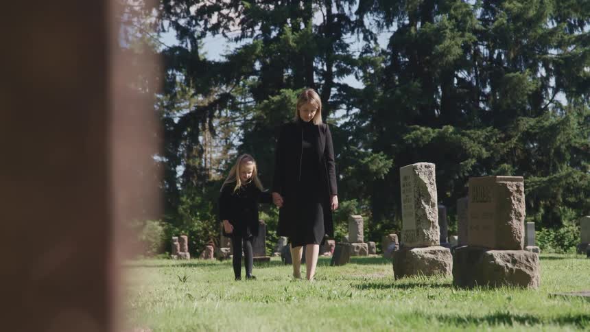 A woman walking and holding a girls hand in a cemetery with trees sky and clouds in the background