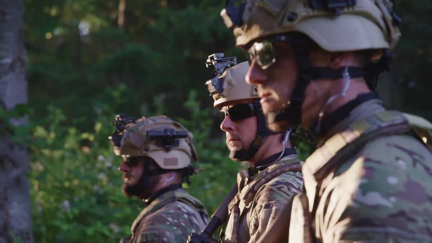 Three soldiers in uniform wearing helmets and sunglasses near a forest at evening
