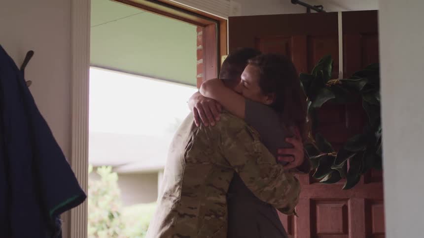 Man wearing military clothing and woman hugging next to a door inside a house