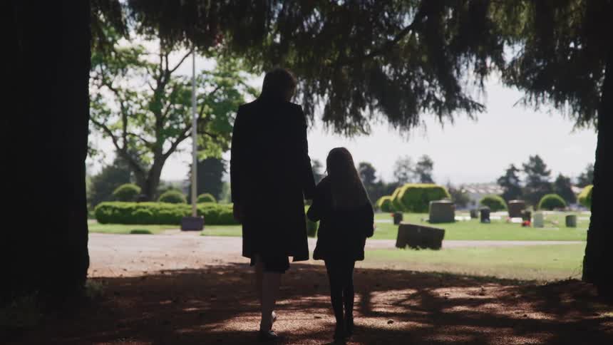 A mother and daughter walk under the shade of a tree holding hands towards the inside of a cemetery at daytime
