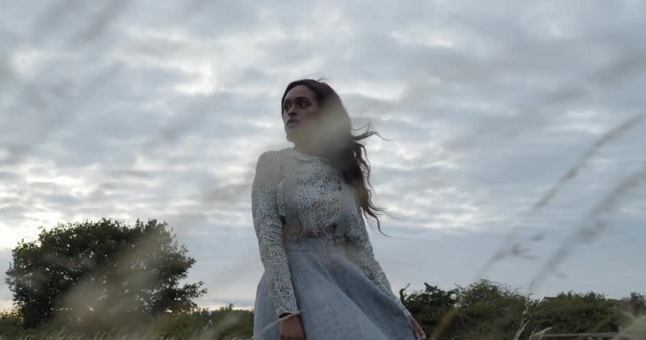 A young woman stands among the tall swaying grass of a field on a cloudy evening