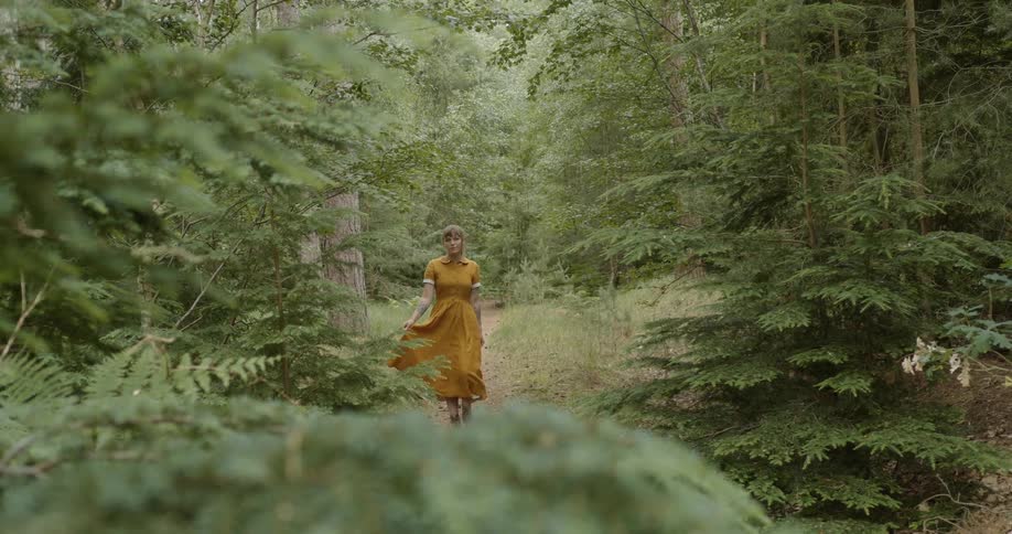 Woman swaying her dress with one hand walks down a path in a forest surround by trees and vegetation at daytime