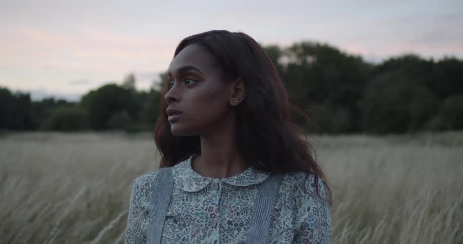 A young woman sits in a field of tall pale grass under an evening sky