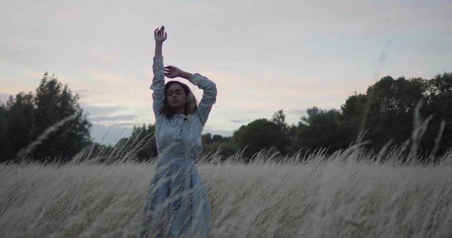 A young woman in an old fashioned dress stretches her arms while standing in a field in the evening