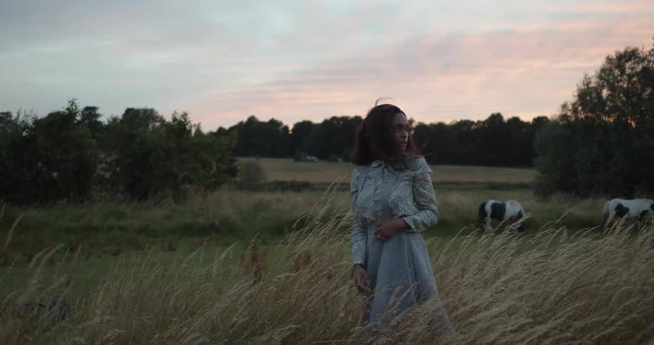 A woman standing in the tall grass pushes her back as horses graze behind her