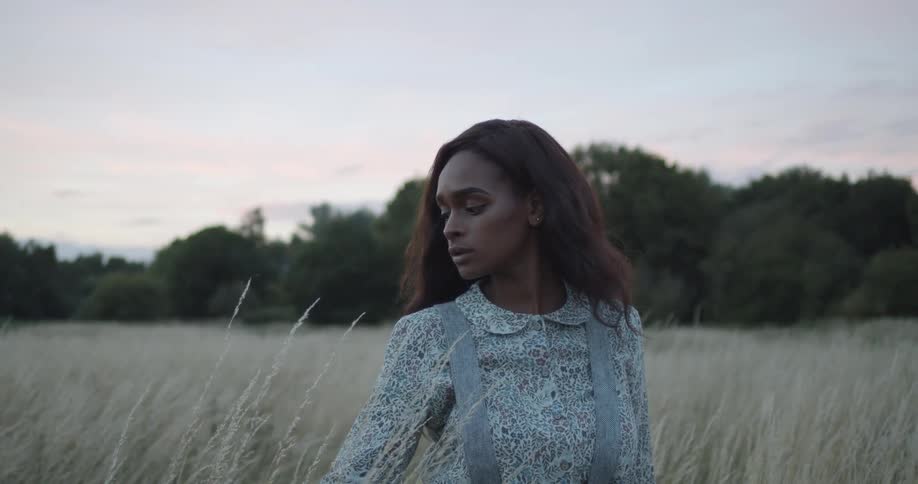 A young black woman standing in a field with long grass with a forest in the distance