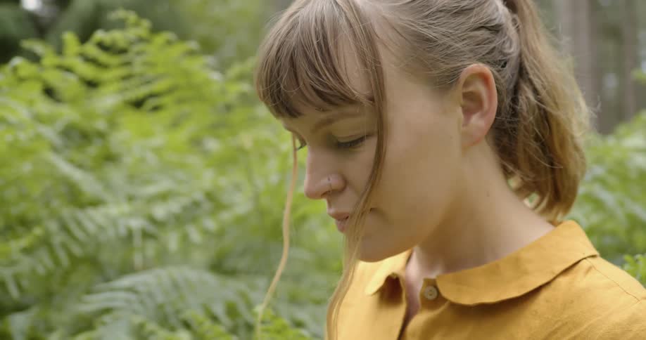 A woman stands near a dense bush in a forest with her head bowed and eyes closed